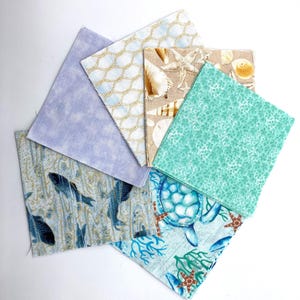 May include: A collection of square fabric pieces with various ocean-themed patterns. Designs include fish, turtles, seashells, and coral in shades of blue, green, and tan. Some fabrics feature a light blue and gold rope design, and a solid light purple fabric.