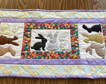 May include: A purple and white quilted table runner with a colorful border and a row of embroidered bunnies in the center. The bunnies are in various colors and patterns, including black, white, brown, and gray.