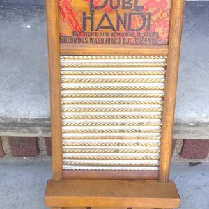 Op de afbeelding: Een vintage houten wasbord met een gegolfd metalen oppervlak. Het wasbord heeft een bord met de tekst "Durl Handi" en "Columbus Washboard Co. Columbus".