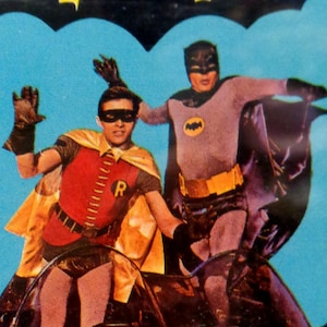Könnte beinhalten: Eine Kassettenhülle mit dem Batman-Logo in Gelb und Schwarz auf blauem Hintergrund. Das Cover zeigt Batman und Robin in einem Fahrzeug mit dem Text: MUSIC CONDUCTED BY NELSON RIDDLE, Featuring BATMAN and ROBIN, plus guest villains: THE PENGUIN, ZELDA THE GREAT, MR. FREEZE, THE RIDDLER, ORIGINAL TELEVISION SOUNDTRACK ALBUM.