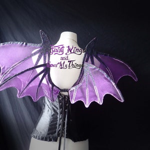 May include: Purple and black bat wings with glitter accents. The wings are attached to a black corset. The text "Fairy Wings and Sparkly Things" is visible on the corset.