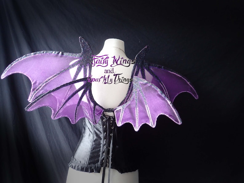 May include: Purple and black bat wings with glitter accents. The wings are attached to a black corset. The text "Fairy Wings and Sparkly Things" is visible on the corset.