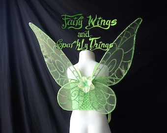 May include: Green fairy wings with a sparkly finish and a flower design. The wings are attached to a white mannequin.