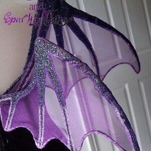 May include: Purple and black fairy wings with glitter accents. The wings are made of sheer fabric and have a pointed shape.