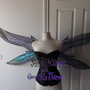 May include: A pair of iridescent dragonfly wings with a black mesh design. The wings are attached to a white mannequin torso wearing a black corset. The text "Fairy Wings and Sparkly Things" is visible in the lower center of the image.