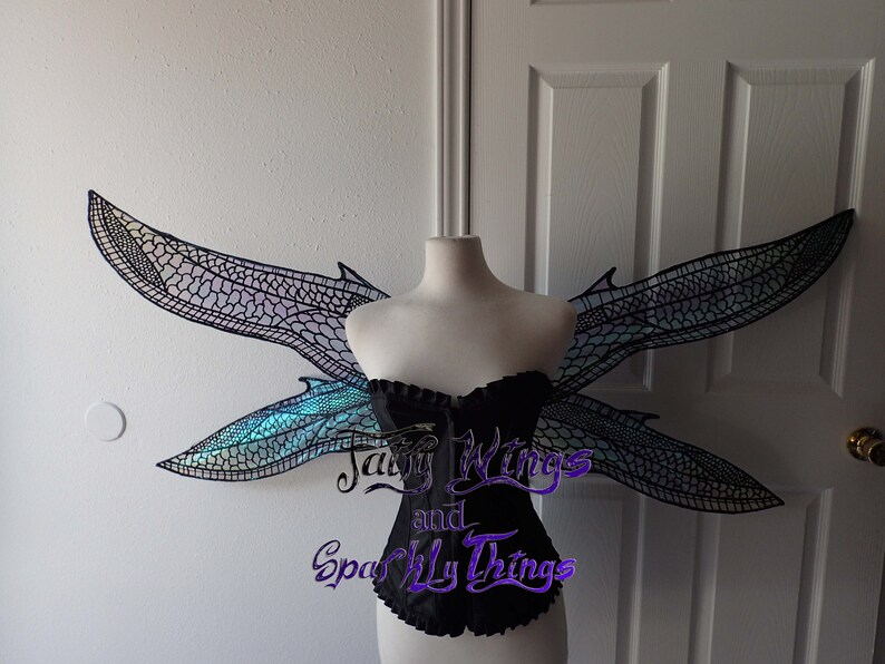 May include: A pair of iridescent dragonfly wings with a black mesh design. The wings are attached to a white mannequin torso wearing a black corset. The text "Fairy Wings and Sparkly Things" is visible in the lower center of the image.