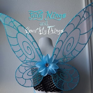 May include: A pair of light blue fairy wings with a white swirl design and glitter accents. The wings are attached to a blue flower with a rhinestone center.