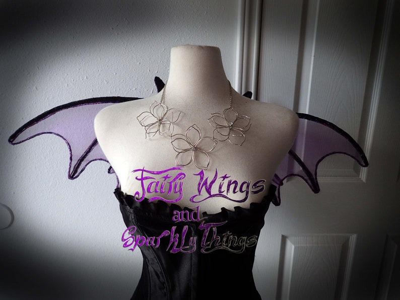 May include: A silver wire flower necklace with three flowers, worn on a mannequin with purple fairy wings. The text "Fairy Wings and Sparkly Things" is visible on the mannequin's bodice.
