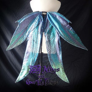 May include: A pair of iridescent blue and green fairy wings with a black mesh pattern. The wings are attached to a black strap and are displayed on a white mannequin.