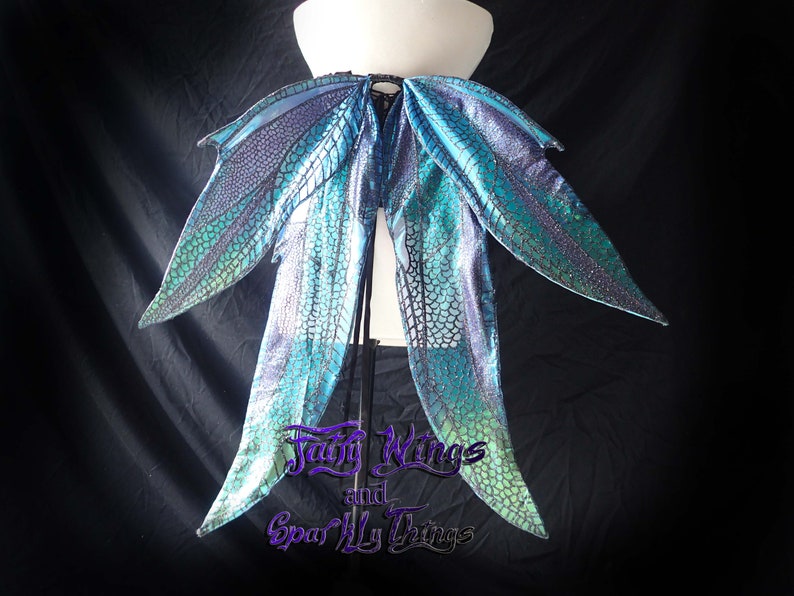 May include: A pair of iridescent blue and green fairy wings with a black mesh pattern. The wings are attached to a black strap and are displayed on a white mannequin.