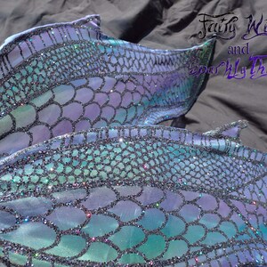 May include: A mermaid tail fin with a blue, green, and purple iridescent scale pattern. The scales are covered in glitter. The tail fin is made of fabric and is designed for swimming.