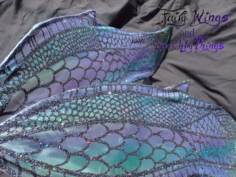 May include: A mermaid tail fin with a blue, green, and purple iridescent scale pattern. The scales are covered in glitter. The tail fin is made of fabric and is designed for swimming.