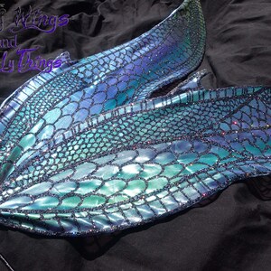May include: A pair of iridescent blue and green fairy wings with a scale pattern and glitter accents. The wings are attached to a black stick.