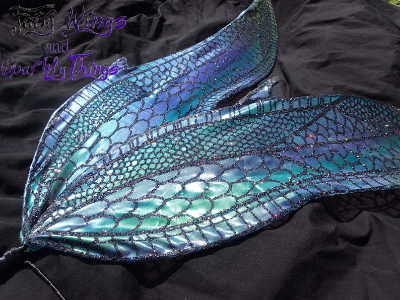 May include: A pair of iridescent blue and green fairy wings with a scale pattern and glitter accents. The wings are attached to a black stick.