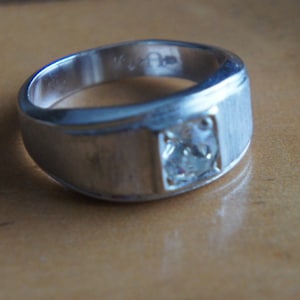 Könnte beinhalten: Ein silberner Ring mit einem einzigen, klaren Edelstein, der in einer quadratischen Fassung sitzt. Der Ring hat ein breites Band und eine polierte Oberfläche.