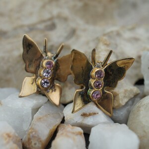 May include: A pair of gold-coloured butterfly stud earrings. Each earring features a butterfly design with three small, light purple gemstones. The earrings are set against a background of white and grey stones.