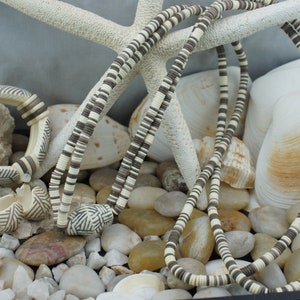 May include: A set of three necklaces made of white and brown ceramic beads. The necklaces are arranged on a bed of white stones and seashells.