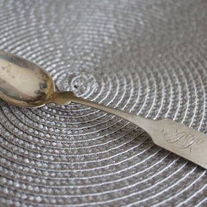 May include: A silver teaspoon with an ornate handle and a monogram on the back. The spoon is lying on a grey, circular placemat.