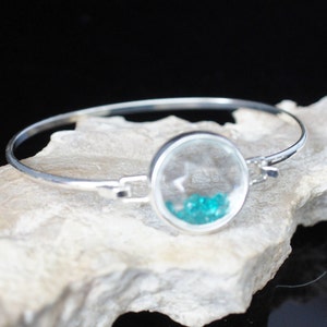 Puede incluir: Un brazalete de plata con un dije redondo de vidrio que contiene pequeñas piedras preciosas turquesa.