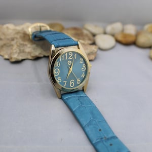 以下が含まれることがあります： 青い革のバンドと白い数字の青い文字盤が付いたゴールドトーンの腕時計。時計はゴールドトーンのバックルが付いています。