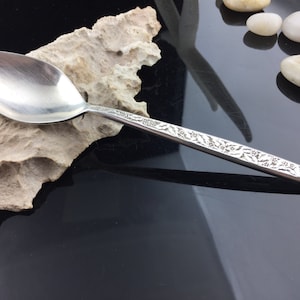 May include: A silver spoon with an ornate floral design on the handle. The spoon is resting on a piece of driftwood.
