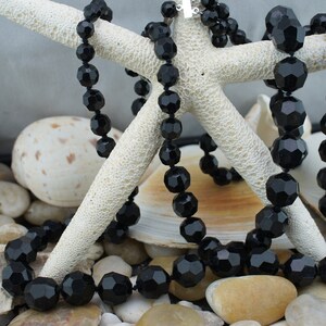 May include: A black beaded necklace made of faceted glass beads. The necklace is draped over a white starfish and seashells.