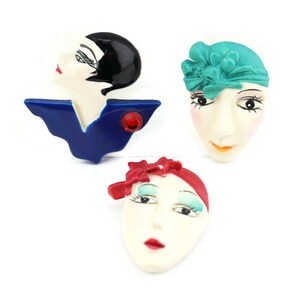 May include: Three vintage ceramic brooches featuring stylized faces. The first brooch is a woman with black hair and a blue dress, the second is a woman with a turquoise hat and the third is a woman with a red headband.