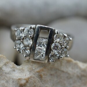 Puede incluir: Un anillo de plata con una piedra central de forma cuadrada y piedras más pequeñas que la rodean.