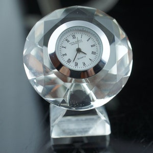 Puede incluir: Un reloj de cristal transparente con una esfera que dice "Fifth Avenue Crystal LTD Quartz". El reloj tiene forma de diamante y una base plateada.