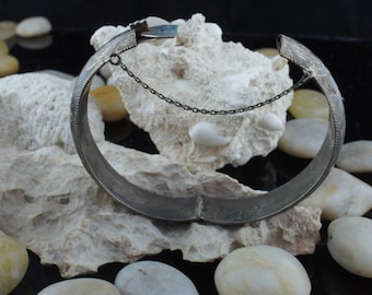 May include: Silver bangle bracelet with an intricate engraved design. The bracelet has a hinged clasp with a small chain.