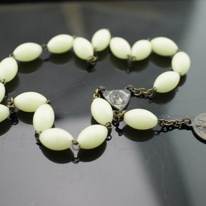 May include: A white glow-in-the-dark rosary with oval beads and a metal pendant featuring a religious image.