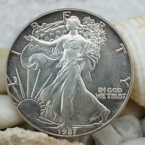 May include: Silver American Eagle coin with a depiction of Lady Liberty walking forward, holding a torch and olive branch. The coin has the inscription "In God We Trust" and the year "1987".
