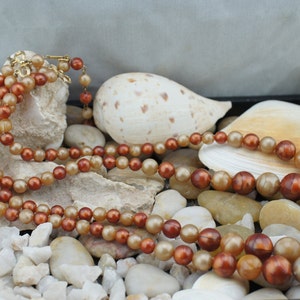 May include: A multi-strand necklace made of brown and beige round beads. The necklace is displayed on a bed of small white stones and seashells.