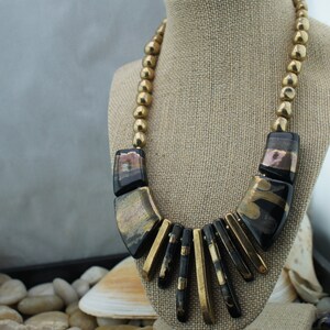May include: A gold and black statement necklace with large, geometric beads. The necklace is made of a combination of round and rectangular beads, with a gold finish and black accents. The necklace is displayed on a mannequin.
