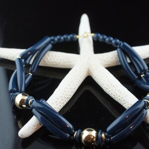 Pode incluir: Um colar azul marinho e dourado com contas grandes e retangulares. O colar é apresentado em uma estrela do mar branca.
