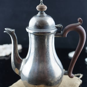 May include: A silver teapot with a wooden handle and a domed lid. The teapot is sitting on a hessian surface.