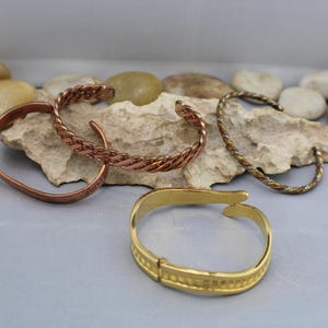 Puede incluir: Cuatro pulseras de metal diferentes, dos son de cobre, una es de oro y una es una pulsera trenzada de cobre y plata. Las pulseras están sobre una superficie de roca marrón claro.