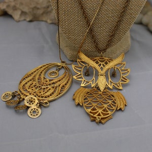 May include: Two gold tone necklaces with intricate filigree designs. One necklace features a large owl pendant with a detailed body and wings. The other necklace has a teardrop-shaped pendant with a dangling chain of small circular charms.
