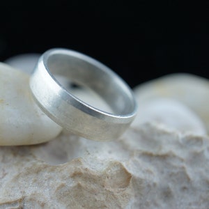 May include: A silver wedding band with a brushed finish. The band is sitting on a white rock.