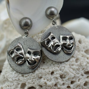 May include: A pair of silver dangle earrings with a textured round disc featuring two silver masks, one smiling and one frowning.