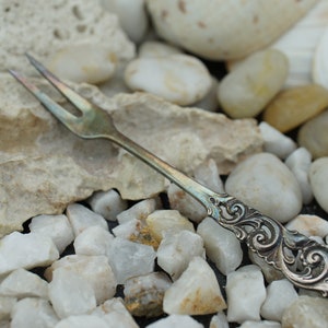 May include: A silver fork with an ornate handle, lying on a bed of white and gray stones.