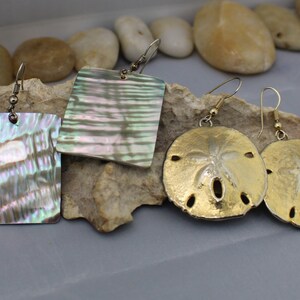 May include: Four pairs of earrings with different designs. Two pairs are square-shaped with iridescent abalone shells. The other two pairs are round, resembling sand dollars, with gold-colored metal. All earrings have hook closures.