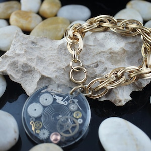 May include: A gold chain bracelet with a clear pendant featuring a circular design of gears and cogs.