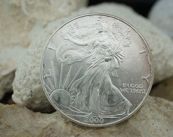 Könnte beinhalten: Eine silberne American Eagle Münze mit dem Bildnis von Lady Liberty, die mit einer Fackel und einem Olivenzweig geht. Die Münze trägt die Inschrift "In God We Trust" und das Jahr "2000".