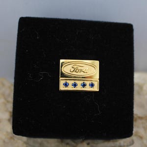 Può includere: Spilla dorata con logo Ford e quattro pietre blu. La spilla è in una scatola di velluto nero.