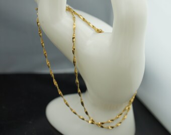 May include: A delicate gold-colored necklace with a twisted rope design. The necklace is draped over a white, hand-shaped display, showcasing its intricate details. The background is a mix of black, white, and gray.