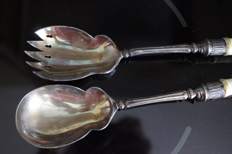 May include: A silver salad serving set with a fork and spoon. The handles are made of a white material with a decorative pattern.
