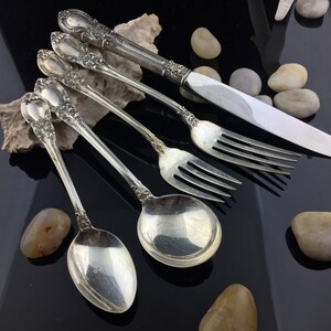 Puede incluir: Un juego de cinco cubiertos de plata plateada, que incluye un cuchillo, un tenedor y tres cucharas. Las piezas tienen un diseño floral adornado.