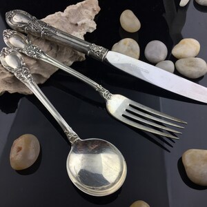 Puede incluir: Un juego de tres cubiertos plateados, que incluye un tenedor, una cuchara y un cuchillo. Los cubiertos tienen mangos adornados con diseños florales.