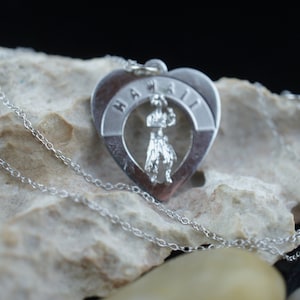 Peut inclure: Un collier avec un pendentif en forme de cœur en argent avec le mot "Hawaii" inscrit sur le bord. Le pendentif présente une découpe d'une danseuse hula au centre.